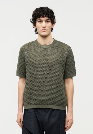 Ung mand med mørkt krøllet hår iført struktureret olivengrøn kortærmet sweater og mørke bukser med striber mod ensfarvet baggrund.