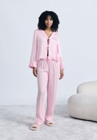 Γυναίκα με σγουρά μαλλιά στέκεται φορώντας ανοιχτό ροζ πιτζάμες και μπεζ παντόφλες δίπλα σε μια λευκή υφασμάτινη πολυθρόνα πάνω σε γκρι χαλί.