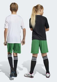 Twee kinderen van achteren in voetbaluniformen: de ene in een wit shirt met groene shorts, de andere in een zwart shirt met groene shorts. Beide dragen zwarte sokken met witte strepen en voetbalschoenen.