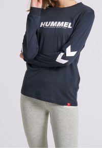 Mörkblå långärmad skjorta med vit "HUMMEL"-logga framtill och chevron-accenter på ärmarna, som parats ihop med grå leggings med en liknande logga.