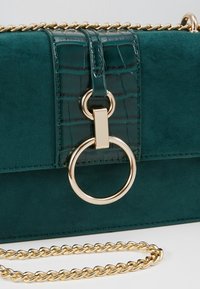 Sac à main vert en daim et cuir imitation crocodile avec bandoulière chaîne dorée et grand fermoir anneau doré sur le rabat avant.