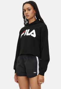 Sudadera recortada negra con logo blanco de FILA; combinada con pantalones cortos negros que tienen rayas laterales blancas y logo de FILA. Tela texturizada, corte relajado.