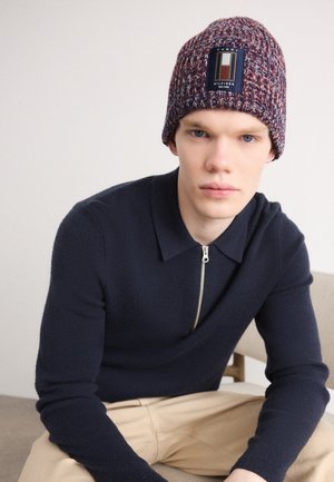 Jeune homme portant un bonnet tricoté multicolore avec un patch logo, un pull à fermeture éclair bleu marine et un pantalon beige, assis devant un fond uni.