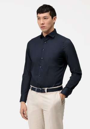 Camicetta blu navy a maniche lunghe con design aderente, bottoni sulla parte anteriore, abbinata a pantaloni beige e una cintura intrecciata blu scuro.