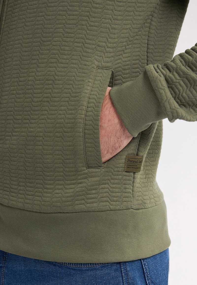 Zielona bluza z kapturem o wzorze, z teksturowaną powierzchnią, z kieszenią kangurką, ściągaczami i metką z logo. Wykonana z miękkiego materiału, w luźnym kroju.