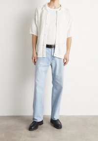 Witte gebreide cardigan met korte mouwen over een wit shirt, gecombineerd met lichtblauwe spijkerbroek en zwarte instappers. Gelaagd patroon op de cardigan.