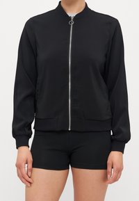 Black bomber jacket in smooth fabric, featuring a high collar, front zipper, and ribbed cuffs, paired with black athletic shorts.