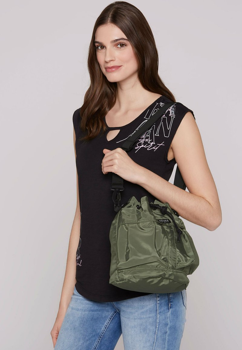 Olive green nylon bucket bag with drawstring closure, featuring adjustable black strap and gathered texture, held by a person wearing a black top.