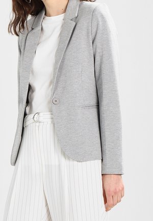 Blazer gris con cuello de pico, cierre de un solo botón y tejido suave. Combinado con pantalones rayados blancos, que muestran un ajuste a medida.