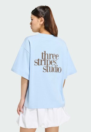 Žena s dlouhými vlasy nosí světle modré oversized tričko s hnědým nápisem "three stripes studio" na zádech a bílou plisovanou sukni.