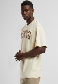 Cream-colored cotton t-shirt featuring "MIAMI" printed in large, brown uppercase letters across the chest. Short sleeves, relaxed fit.