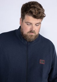 Navy zip-up jacket with a ribbed texture, high collar, and a small red logo patch on the left chest area. Zipper front closure.