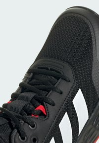 Černá sportovní bota s síťovaným svrškem, na kterém jsou tři bílé pruhy, červený akcent a černá texturovaná podrážka. Šněrování je černé s oky.