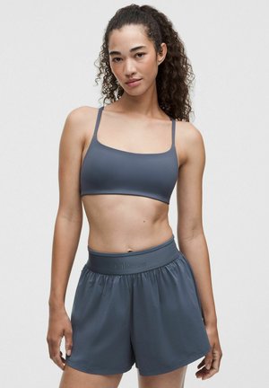 Kvinde med krøllet hår iført en grå sports-bh og grå Lululemon-shorts, stående foran en ensfarvet lys baggrund.