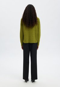 Žalias megztinis su laisvu siluetu, derinamas su juodomis kirpimo kelnėmis ir aštriais juodais aukštakulniais, matomas iš nugaros.