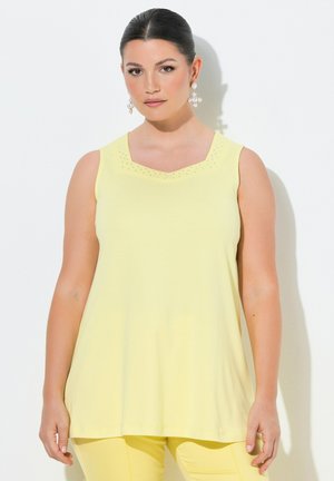 Kobieta o ciemnych włosach, ubrana w żółty top bez rękawów i pasujące spodnie, nosząca duże perłowe kolczyki w kształcie krzyża, stojąca na białym tle.