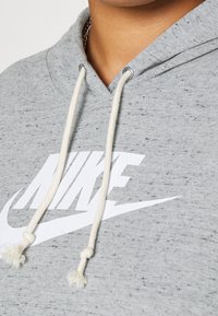 Grå hoodie gjord av mjukt tyg, med en stor vit Nike-logga, knytband med tofsar och en prickig textur.