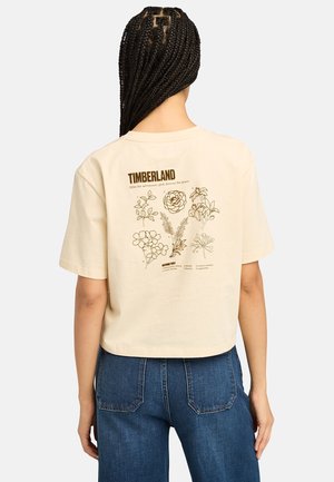 Persoon met gevlochten haar die een beige Timberland T-shirt draagt met botanische bloemillustraties en namen op de achterkant, gecombineerd met een blauwe spijkerbroek.