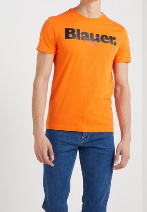 Mann trägt ein leuchtend orangefarbenes kurzärmeliges T-Shirt mit dem Text "Blauer USA" und blaue Jeans, steht vor einem schlichten hellen Hintergrund.