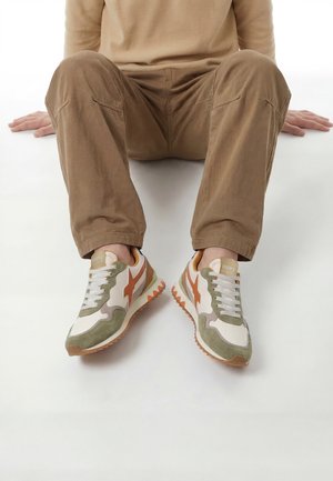 Person sitzt mit angewinkelten Beinen, trägt beige Hose und weiße, grüne und orange Freizeit-Sneaker mit Schnürsenkeln auf weißem Hintergrund.