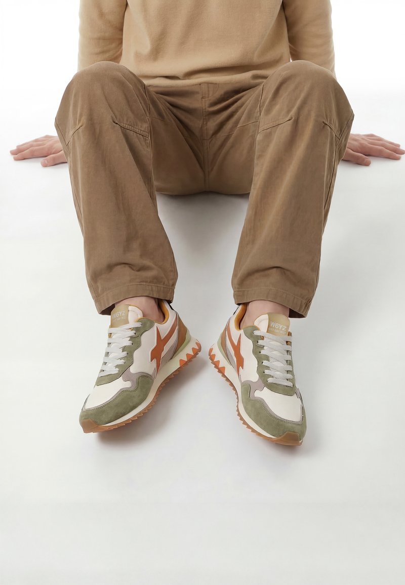 Personne assise, jambes pliées, portant un pantalon beige et des baskets décontractées blanches, vertes et orange avec des lacets sur fond blanc.