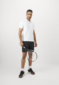 Balta polo marškiniai trumpomis rankovėmis, juodos šortai ir juodi sportiniai bateliai, laikantis teniso raketę su raštuotu tinklu.