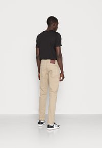Calças beige de corte slim com um bolso traseiro e etiqueta bordô, combinadas com uma camisa preta de mangas curtas e ténis preto e branco.