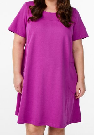 Woman wearing a loose-fitting, short-sleeve magenta dress with a round neckline and a pocket on the right side.