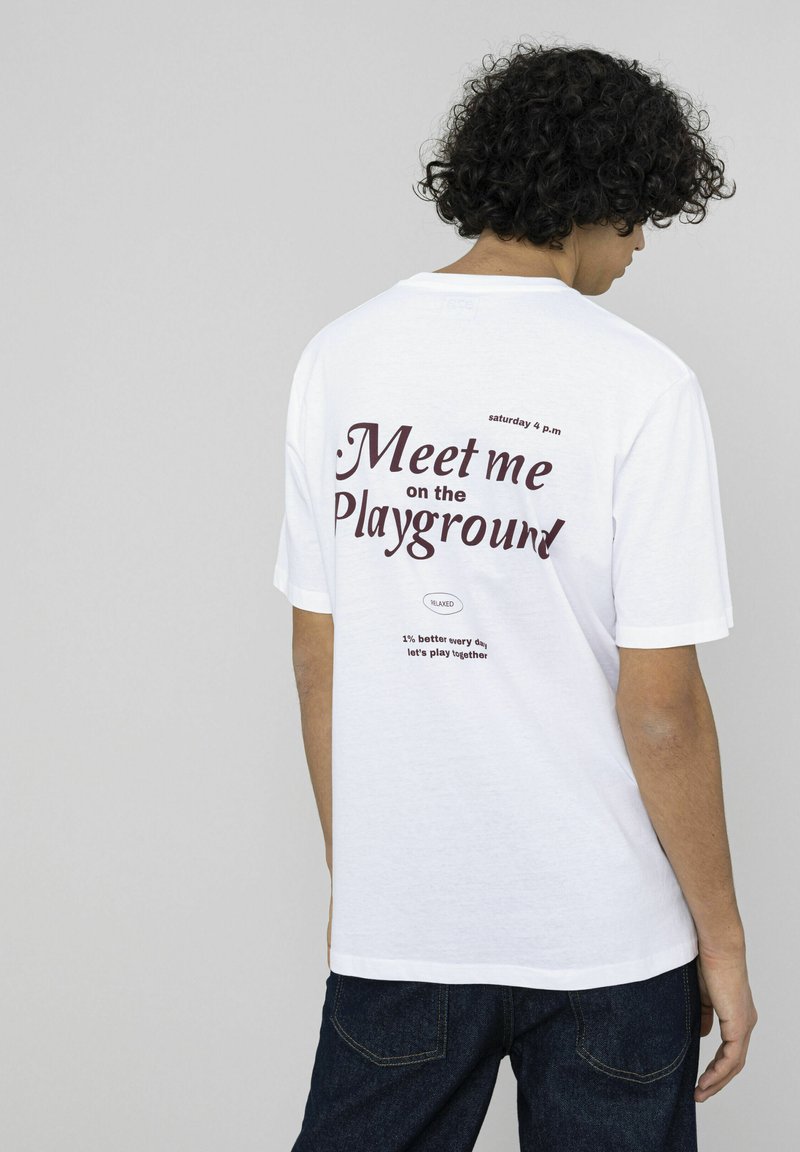 T-shirt en coton blanc avec un imprimé graphique au dos. Le texte comprend "Rendez-vous sur le terrain de jeu" en bordeaux et d'autres phrases en dessous.
