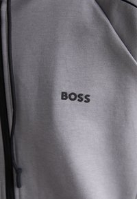 Veste zippée grise en tissu doux, avec une texture lisse et un logo noir "BOSS" sur la poitrine. Comprend des accents de fermeture éclair noirs.