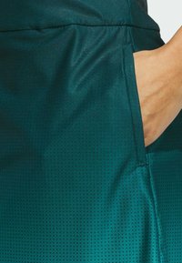 Ciemnoturkusowa tkanina sportowa o gładkiej fakturze z wzorem kropek oraz boczną kieszenią z widocznym przeszyciem.