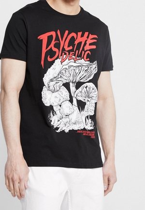 Mężczyzna w czarnej koszulce z dużym, białym, psychodelicznym nadrukiem grzyba oraz wyrazistym czerwonym napisem „PSYCHEDELIC” na przodzie.