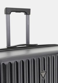 Valise rigide noire avec texture rayée verticale, poignée télescopique grise et détail de logo. Comprend une fermeture éclair et des accents robustes.