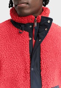 Czerwona, strukturalna kurtka polarowa z wysokim kołnierzem, granatowymi akcentami, zamkiem błyskawicznym z przodu oraz zatrzaskami; wyposażona w sznurek do regulacji dopasowania.