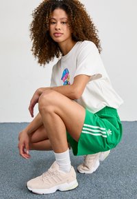 White graphic t-shirt with colorful cartoon design; green shorts with white three-stripe pattern; light pink sneakers; white ankle socks.