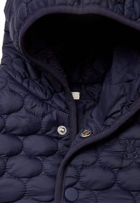 Granatowa, pikowana kamizelka z kapturem, zatrzaskami i subtelnym haftem. Materiał ma gładką, błyszczącą teksturę i puszysty fason.