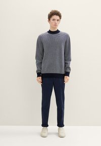 Marine gestreepte textuurtrui met lange mouwen en opgerolde manchetten, gecombineerd met donkerblauwe rechte broek en witte sneakers. Effen achtergrond.