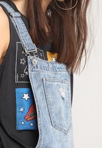 Jeansbyxor med justerbara axelremmar, ljusblå färg, slitna detaljer, framficka; bärs över en svart grafisk linne med färgglada mönster.