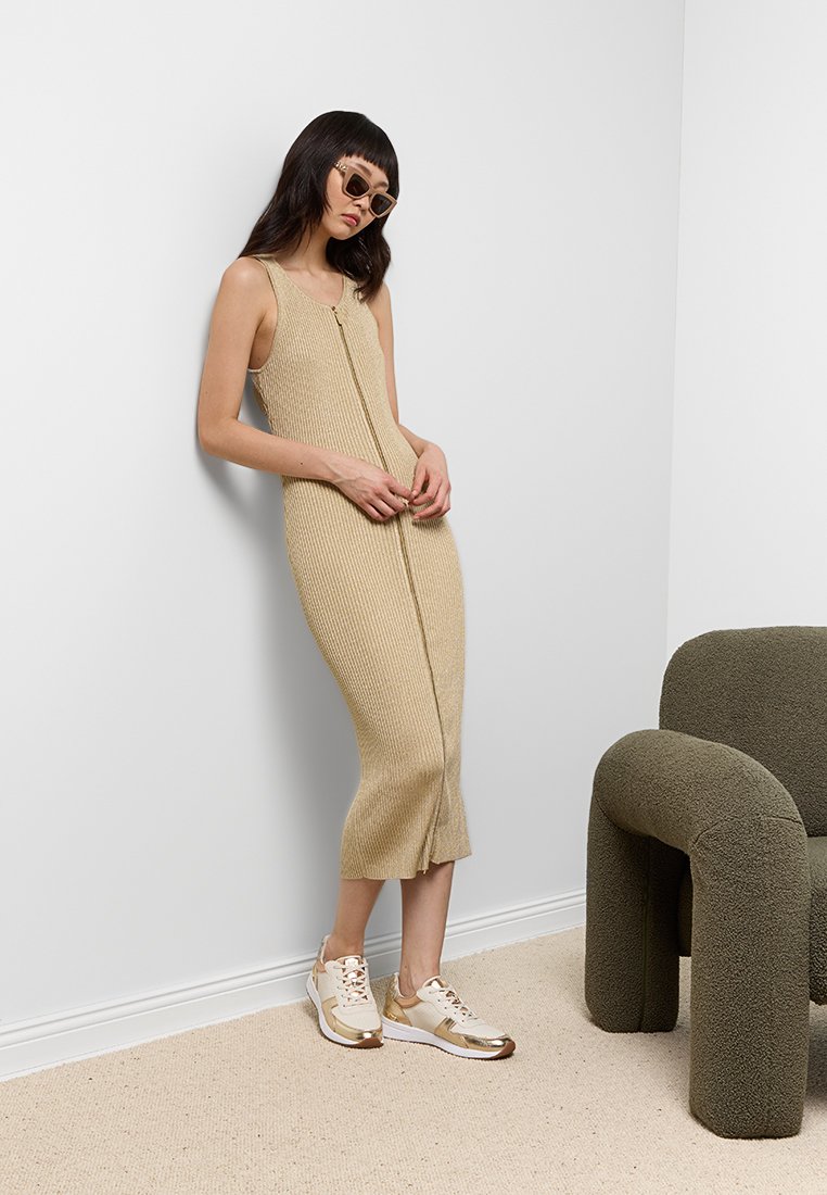 Robe sans manches beige à côtes avec une fermeture éclair à l'avant, portée avec des baskets trimées de métal doré. Le mannequin s'appuie contre un mur clair.