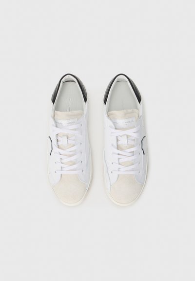 Białe sneakersy z czarnymi akcentami na pięcie, zamszowym czubkiem i teksturowanymi bokami; posiadają białe sznurowadła oraz wyściełany język.