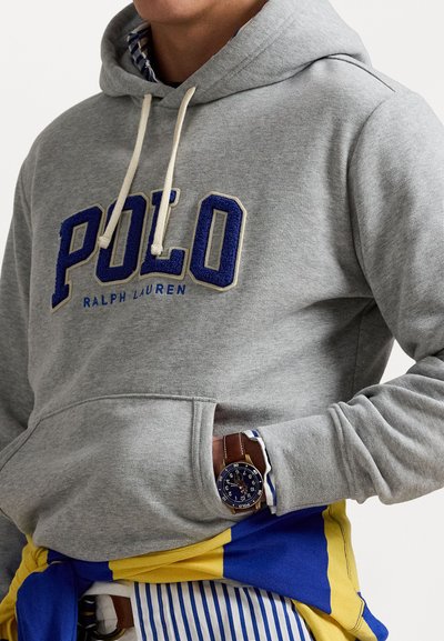 Sivá mikina s textúrovaným modrým logom "POLO", so šnúrkami a prednou vreckom. Nosená cez modro-žltý prúžkovaný tričko s hodinkami.