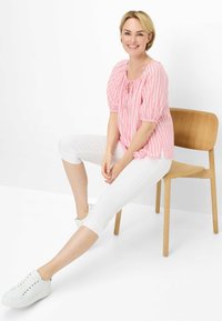 Roze en wit gestreepte blouse met pofmouwen en strikdetail, gecombineerd met witte cropped broek en witte sneakers, zittend op een houten stoel.