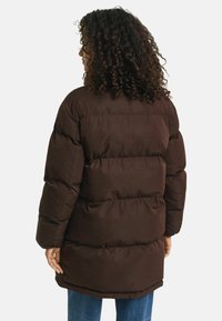 Bruine pufferjas met een lange silhouette, gestalten in een doorgestikte ontwerp en dikke vulling. Kenmerkt een hoge kraag en een relaxte pasvorm. Mouwen gevoerd met fleece.