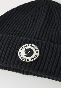 Juodas megztas kepurė su raukšlėta tekstūra ir apvaliu baltu logotipu, kuriame pavaizduota stilizuota lapė ir tekstas "FJÄLLRÄVEN SWEDEN 1960."