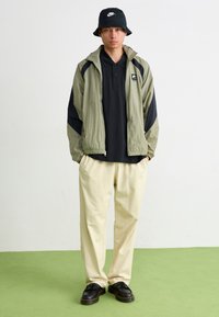 Λαδί πράσινο μπουφάν με μαύρες λεπτομέρειες, μαύρο πόλο μπλουζάκι, κρέμα παντελόνι και μαύρες παπούτσια. Καπέλο bucket με λευκό λογότυπο.