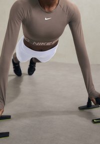 Person in braunem Langarmoberteil und weißen Shorts hält Push-Up-Stangen, während er eine Plank-Übung auf einem grauen Boden macht.