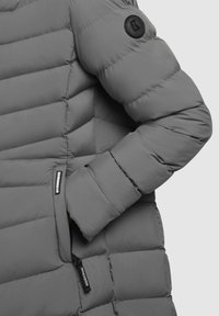 Close-up of a gray quilted jacket sleeve with hand in side pocket, featuring circular black logo patch and zipper pull tabs.
