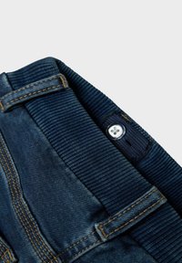 Ceinture en denim bleu foncé avec texture côtelée. Présente des coutures jaunes et un bouton en argent. Insertion élastique pour un ajustement réglable.