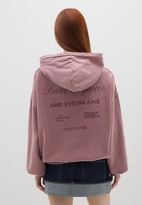 Felpa rosa over-size con cappuccio, con testo stampato sul retro. Abbinata a una minigonna in denim, che mostra una combinazione di due tonalità.