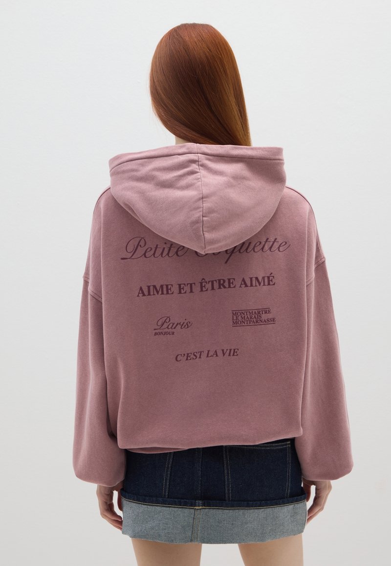 Felpa rosa over-size con cappuccio, con testo stampato sul retro. Abbinata a una minigonna in denim, che mostra una combinazione di due tonalità.